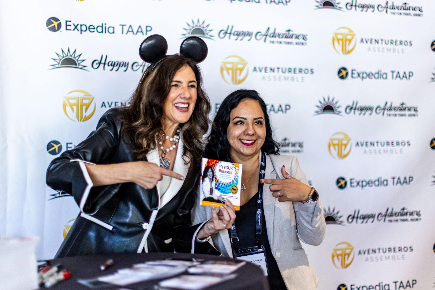 Two women smile and pose together at an event, with one wearing mouse ears and both pointing at a small bag of candy. Event banners with logos are visible in the background.