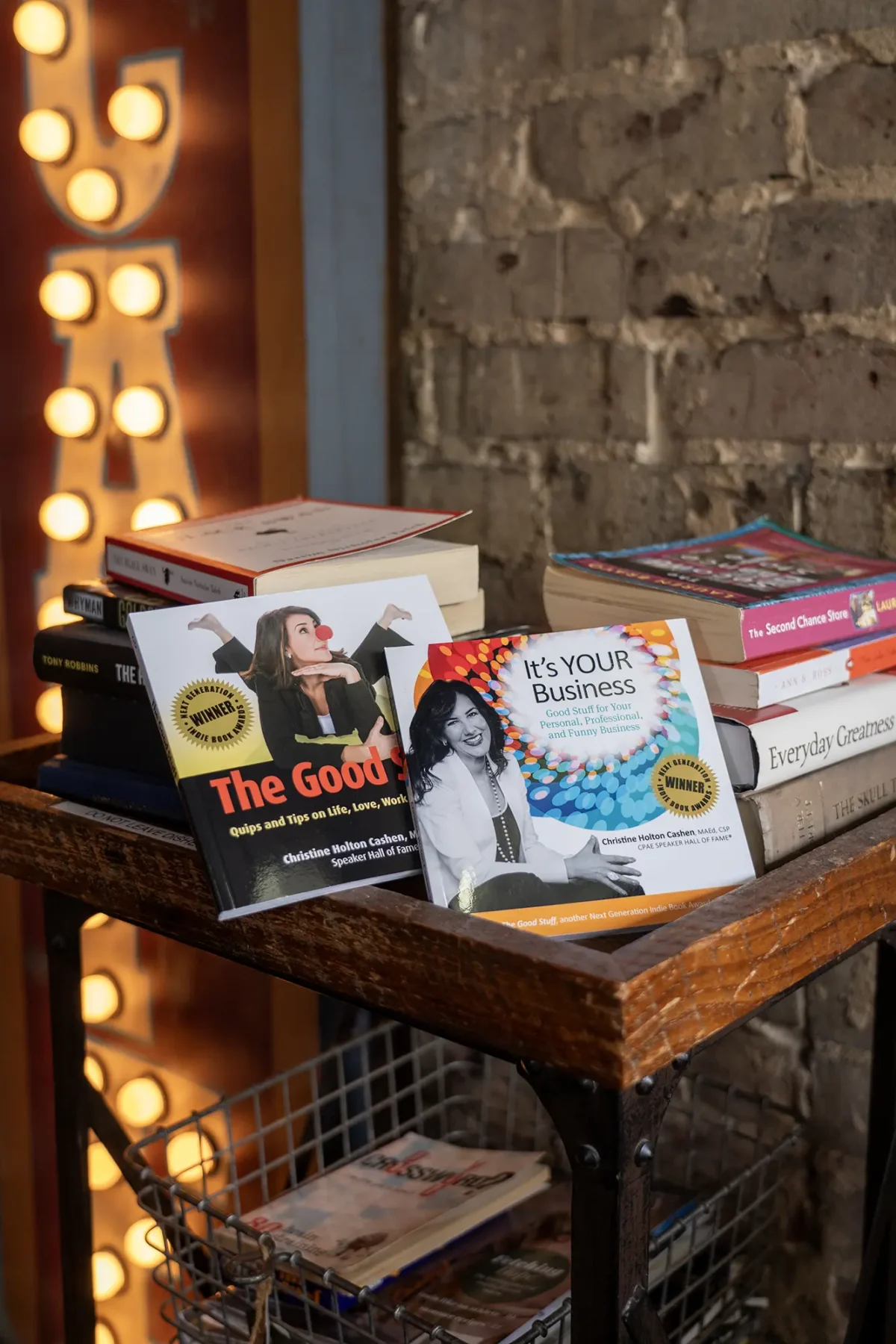 A wooden cart holds several books, including “The Good Girl’s Guide to Being a Boss” and “It’s YOUR Business.” Other books are stacked nearby, all set against a rustic brick wall and a lit retro-style lamp.