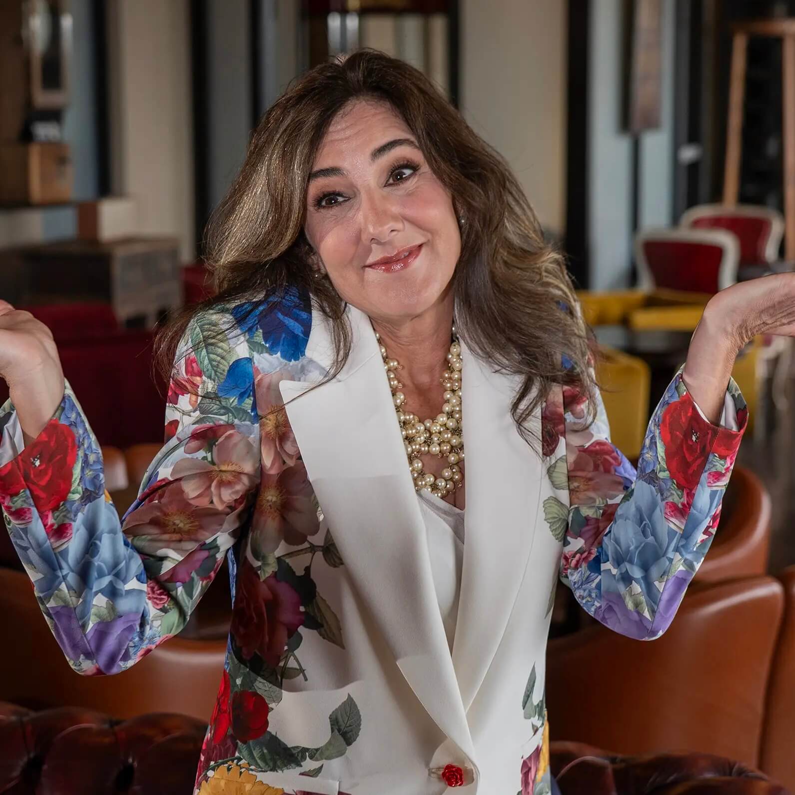 A woman wearing a floral blazer and pearl necklace stands indoors, smiling and shrugging with her palms up, expressing a playful or uncertain gesture.
