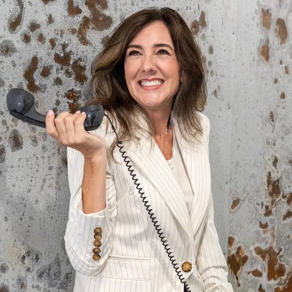 A woman with wavy brown hair, wearing a white pinstripe blazer, smiles while holding a black corded telephone receiver, standing against a textured, speckled wall.