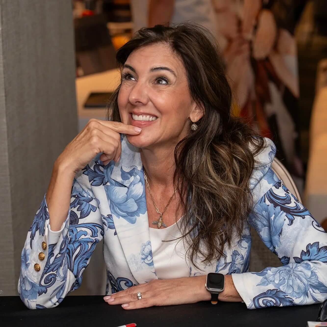 A smiling woman with long brown hair sits at a table, resting her chin on her hand. She wears a blue floral blazer, a white top, and a smartwatch, looking upward with a thoughtful expression.