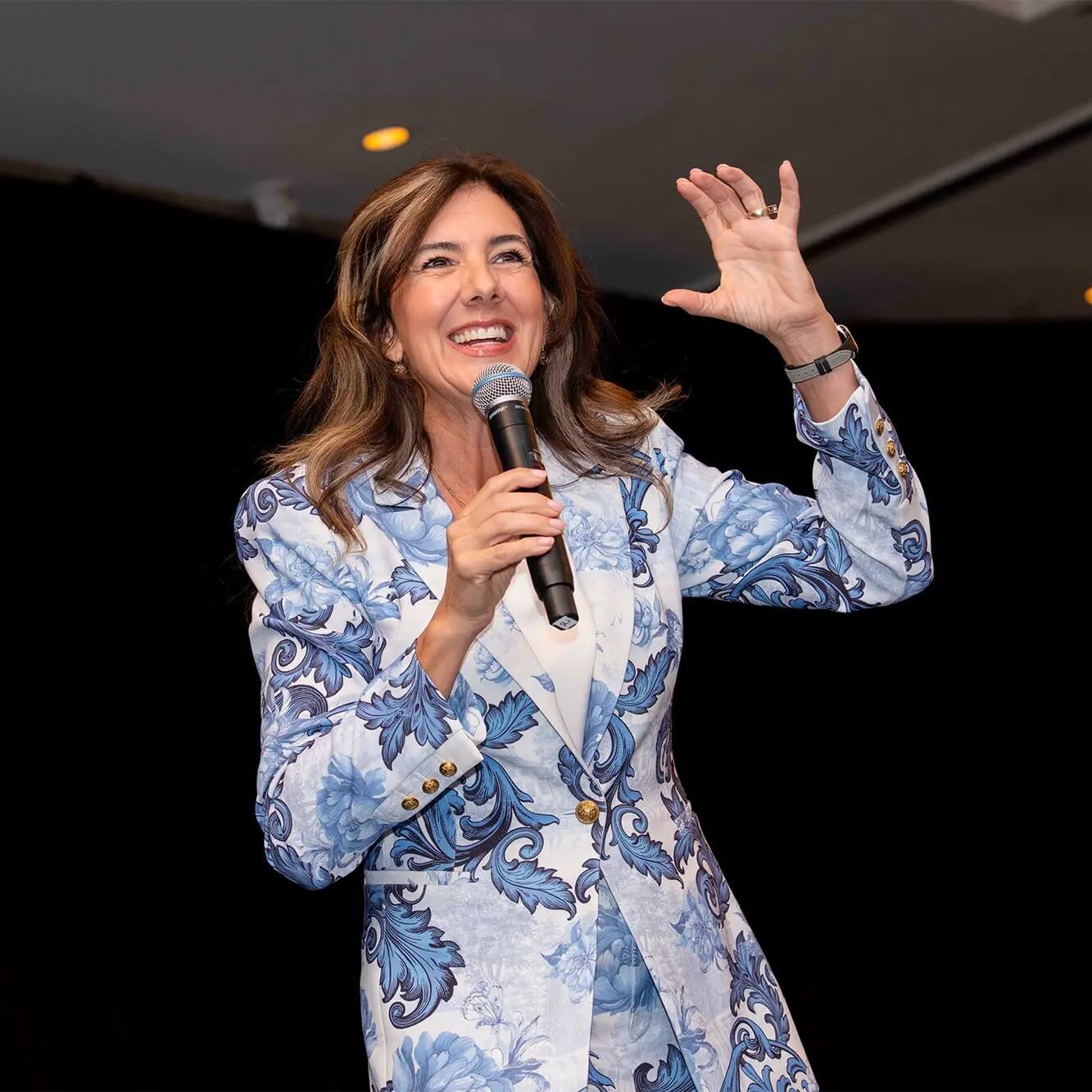 A woman in a blue and white floral suit speaks enthusiastically into a microphone, smiling and gesturing with one hand, against a dark background.