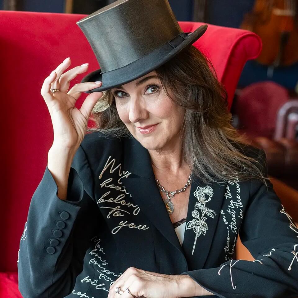 A smiling woman with wavy brown hair tips a black top hat. She wears a dark blazer decorated with white cursive writing and rose illustrations, sitting in front of a bright red chair.