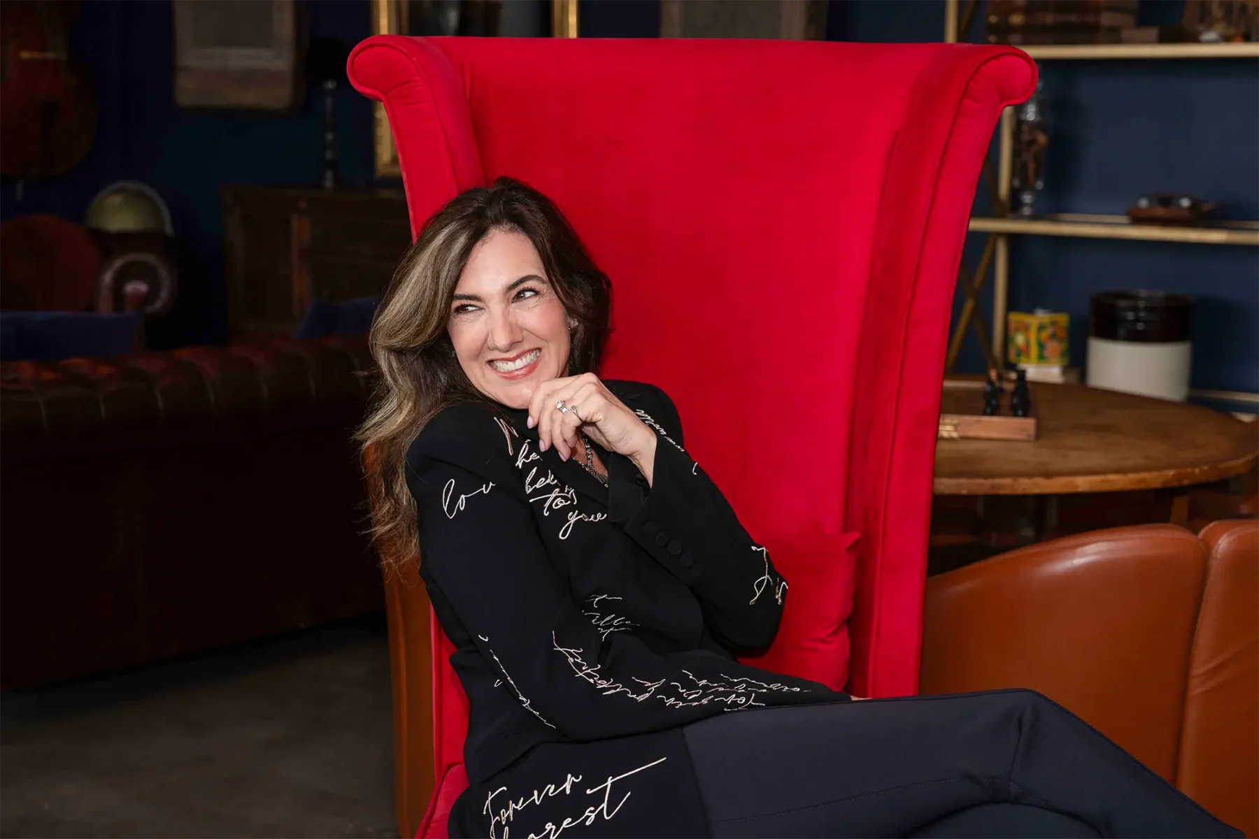 A woman with wavy brown hair sits smiling in a large, bright red chair. She wears a black suit with white script writing and rests her chin on her hand in a relaxed, cheerful pose. The background includes shelves and brown furniture.