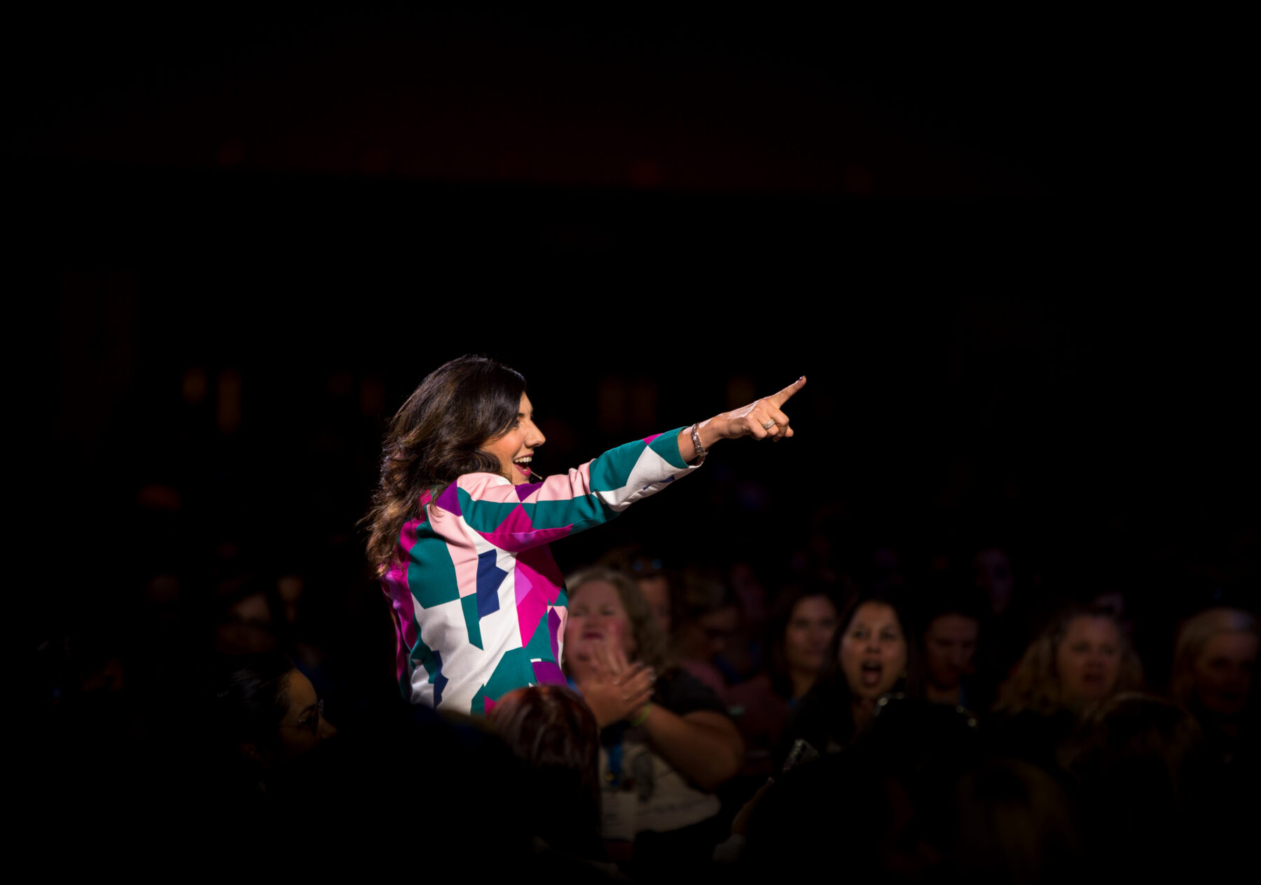 A woman wearing a colorful geometric-patterned jacket stands and points enthusiastically toward the audience, who are watching her with interest in a dimly lit room.