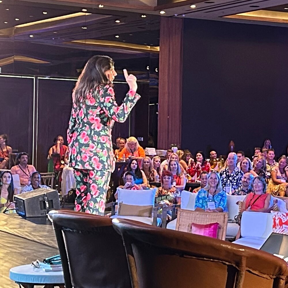 A woman in a colorful floral suit stands on stage, gesturing while speaking to a large seated audience in a well-lit event hall. The audience appears engaged and attentive.