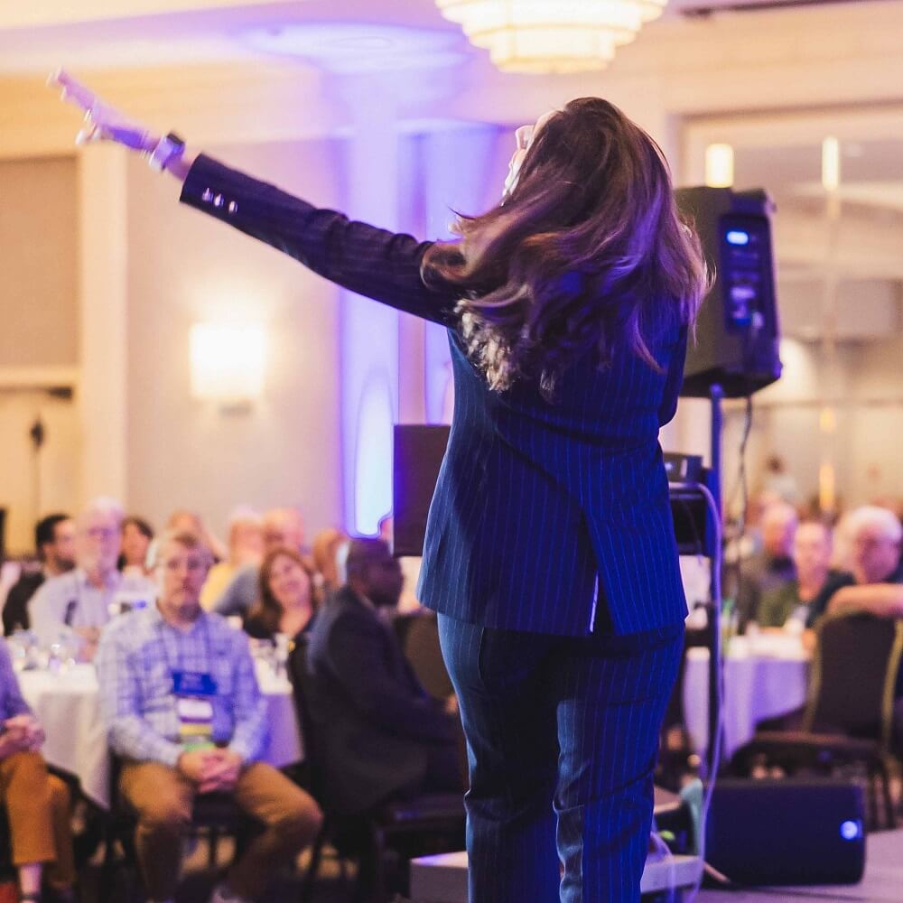 A person in a blue pinstripe suit stands on stage facing an audience, pointing upward with one hand. The audience sits at round tables in a well-lit banquet hall.