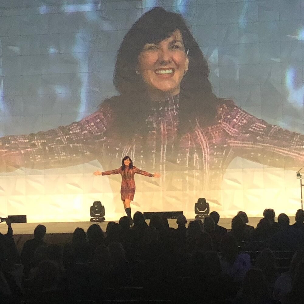 A woman stands on stage with arms outstretched, smiling, in front of a large projected image of herself. Stage lights illuminate her while the audience watches in the foreground.