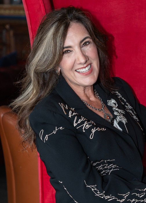 A woman with long brown hair and highlights, wearing a black blazer with white script text, smiles while seated against a red background.