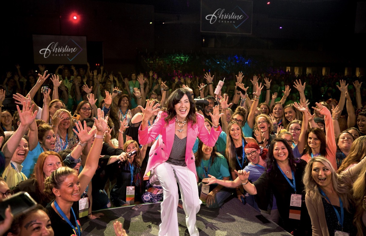 A woman in a pink blazer and white pants stands smiling on stage, surrounded by a large, joyful crowd of people raising their hands at an indoor event. Signs reading Christine Caine are visible in the background.