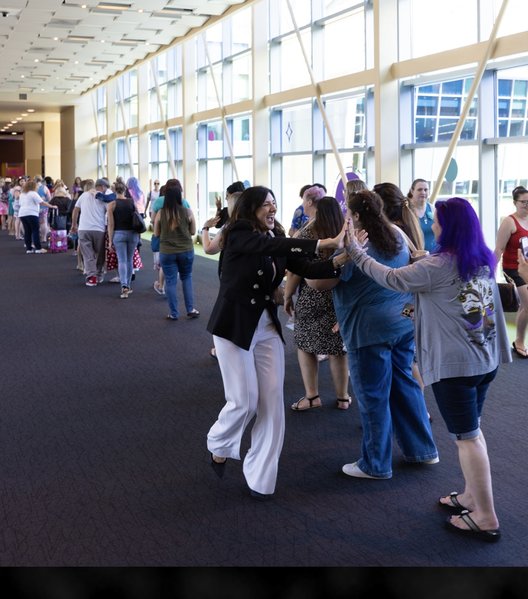 A woman enthusiastically high-fives a line of people in a brightly lit hallway with large windows, as others look on and smile, creating a lively, welcoming atmosphere.