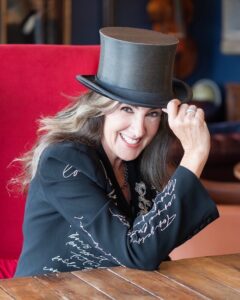 A smiling woman with long wavy hair tips a tall black top hat while sitting at a wooden table. She wears a black jacket with white script writing and a red chair is visible behind her.